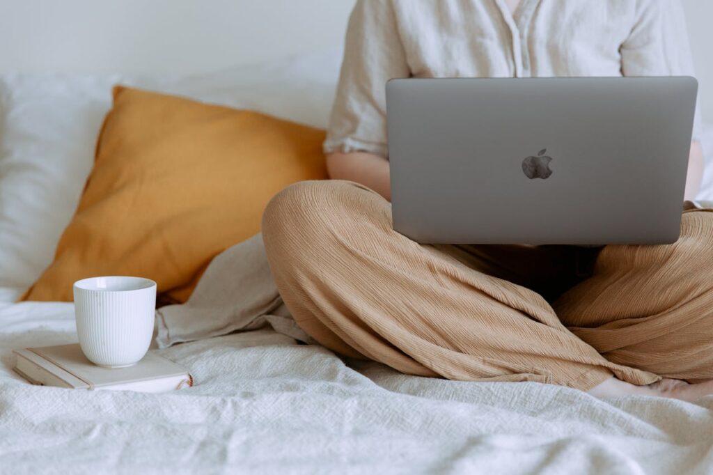 Utbildning Distans Online Bild med ansiktslös kvinna i avslappnad outfit som sitter på sängen med benen i kors, håller en laptop i knäet och utbildar sig på distans hemifrån